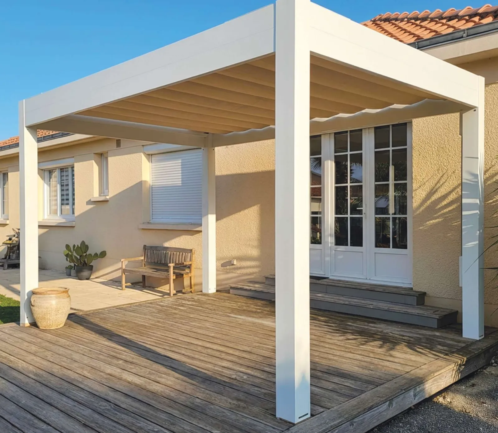 Pergola en aluminium blanc sur une terrasse en bois, devant une maison de plain-pied aux tuiles canal
