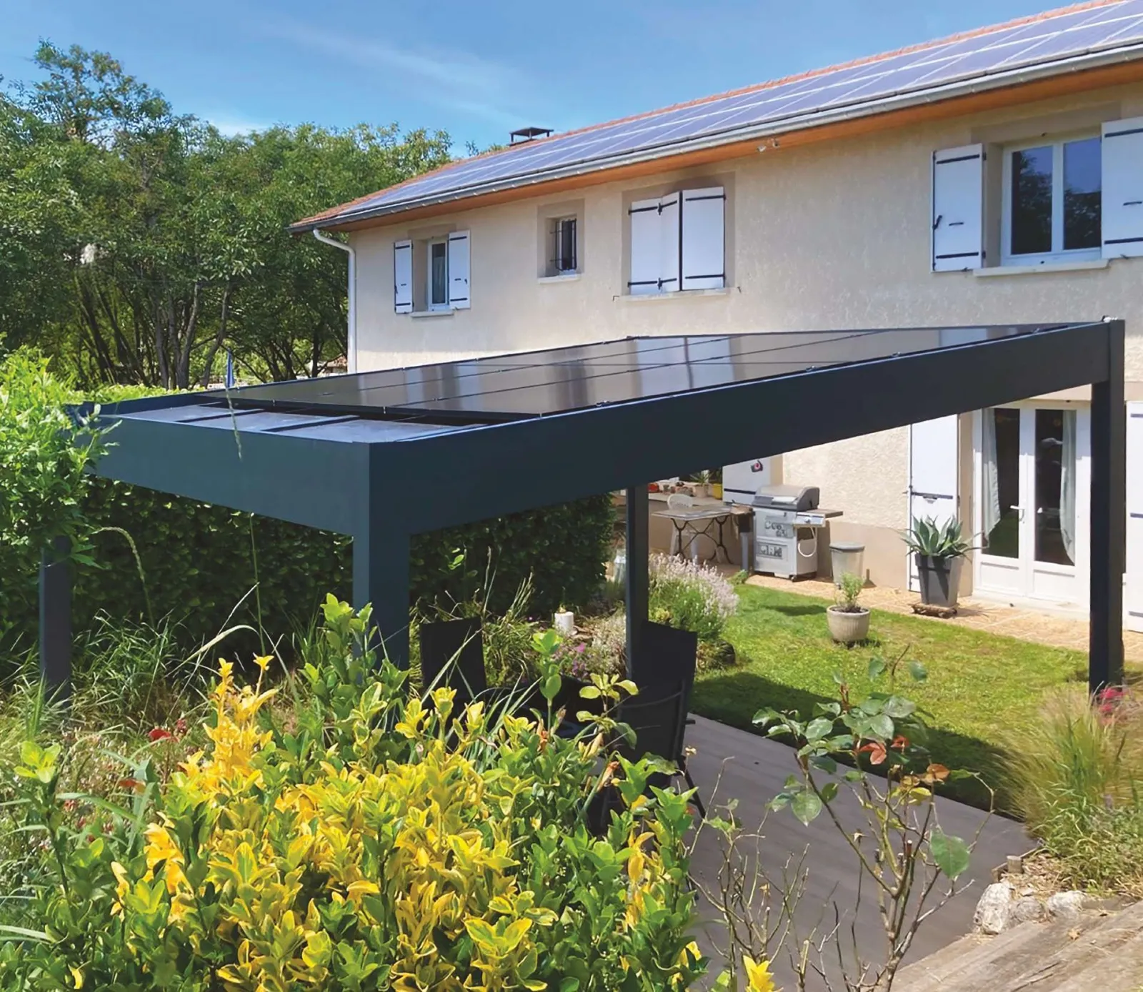 Maison au toit équipé de panneaux photovoltaïques, pergola solaire en aluminium anthracite au premier plan, jardin fleuri en plein soleil